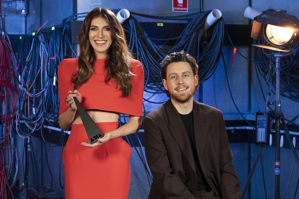 Comedians Melanie Bracewell in a red outfit holding an ARIA Award and Tim McDonald in a brown suit posing backstage amid studio cables